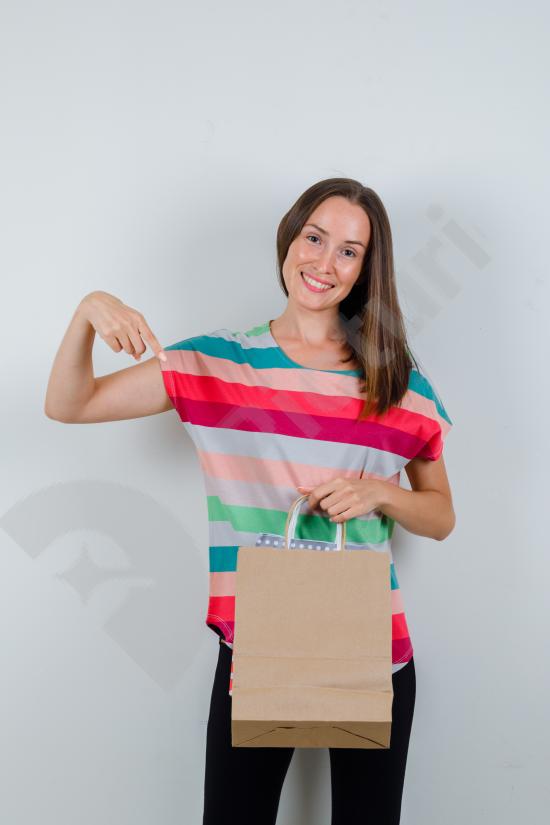 Front view of a young, smiling woman wearing a casual t-shirt and pants, pointing at paper bags. Ideal for shopping and retail themes