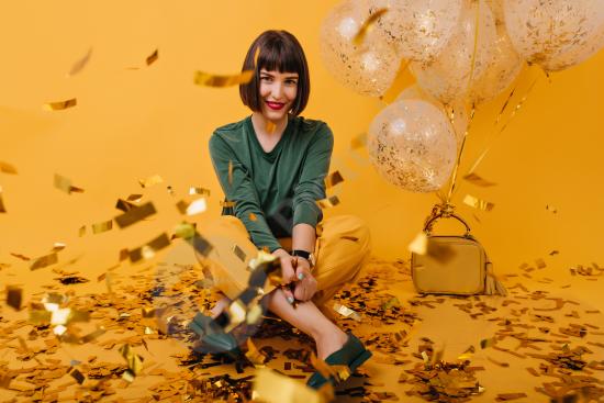 A cheerful brunette girl in a green sweater sitting in confetti, smiling happily in an indoor setting. Perfect for celebrations New Year and joyful mo...