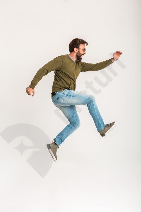 Handsome bearded man jumping and running in stylish casual outfit with sweatshirt, jeans, and sunglasses, isolated on white background.
