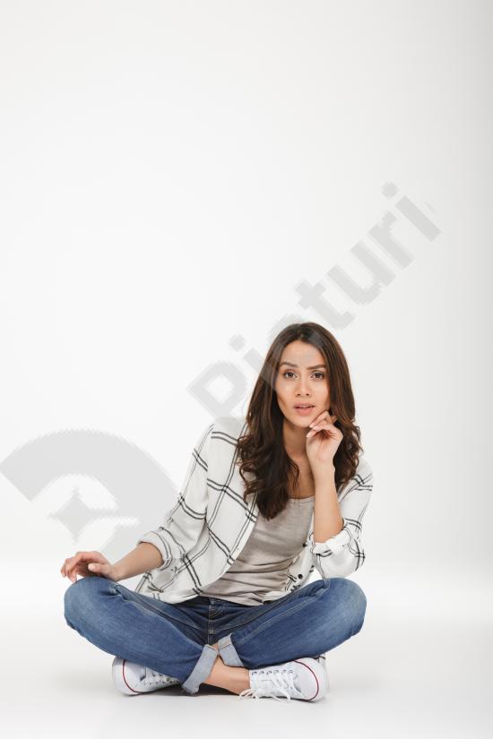 Vertical portrait of a calm brunette woman wearing a shirt, sitting on the floor and looking at the camera against a gray background.