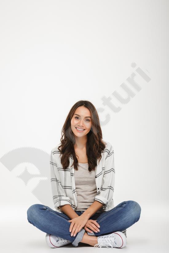 Vertical portrait of a smiling brunette woman wearing a shirt, sitting on the floor and looking at the camera against a gray background.