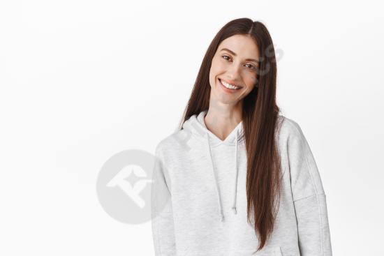 Beautiful young woman with long dark hair smiling gently, tilting her head while wearing a hoodie, standing against a white wall in a candid lifestyle...