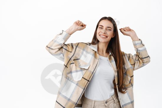 Happy stylish woman dancing with hands up and smiling, celebrating something while standing on a white background, expressing joy and positive energy.