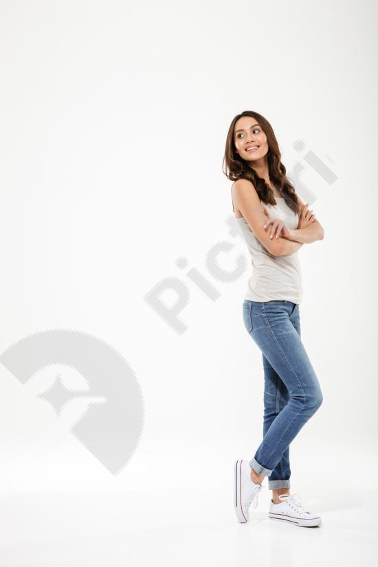 Full-length portrait of a pleased brunette woman posing sideways with crossed arms, looking back. Confident expression on a neutral gray background.